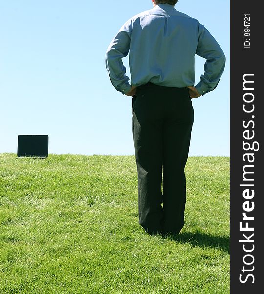 Business man in blue shirt standing holding his hands on his waist in the grass looking towards laptop that's in the grass. Business man in blue shirt standing holding his hands on his waist in the grass looking towards laptop that's in the grass