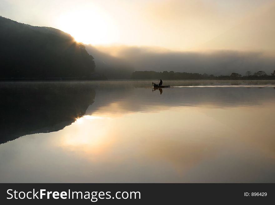 A lone fisherman on a tranquil and misty lake with the sun rising in the background. A lone fisherman on a tranquil and misty lake with the sun rising in the background.