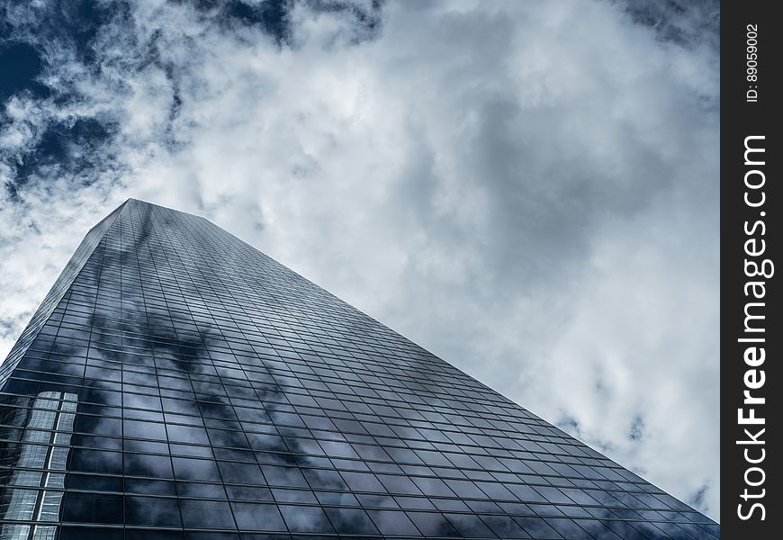 View pointing upwards towards the top of a skyscraper, constructed of mainly steel and glass with light filtering through clouds casting a shadowy pattern on the facade of the building. View pointing upwards towards the top of a skyscraper, constructed of mainly steel and glass with light filtering through clouds casting a shadowy pattern on the facade of the building.