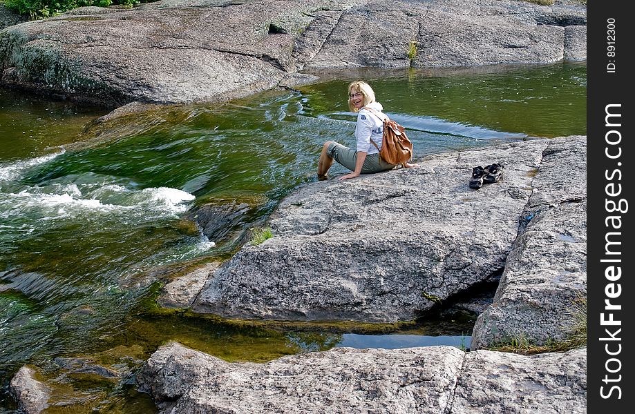 A happy woman is sitting on big stone immersing legs into rapid stream in a very picturesque place. A happy woman is sitting on big stone immersing legs into rapid stream in a very picturesque place.