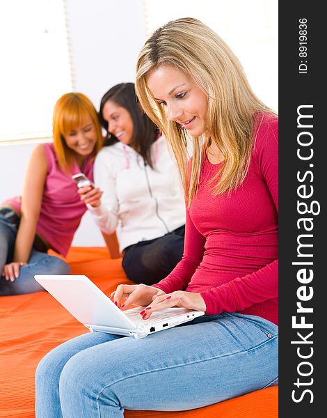 Three young women sitting on bed. One of them working on laptop. Focused on blonde girl with laptop. Three young women sitting on bed. One of them working on laptop. Focused on blonde girl with laptop.