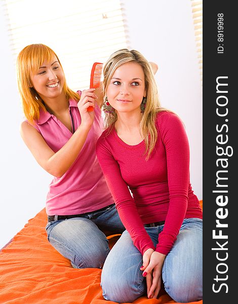 Red-haired woman combing her friend. They're sitting on bed. Front view. Red-haired woman combing her friend. They're sitting on bed. Front view.