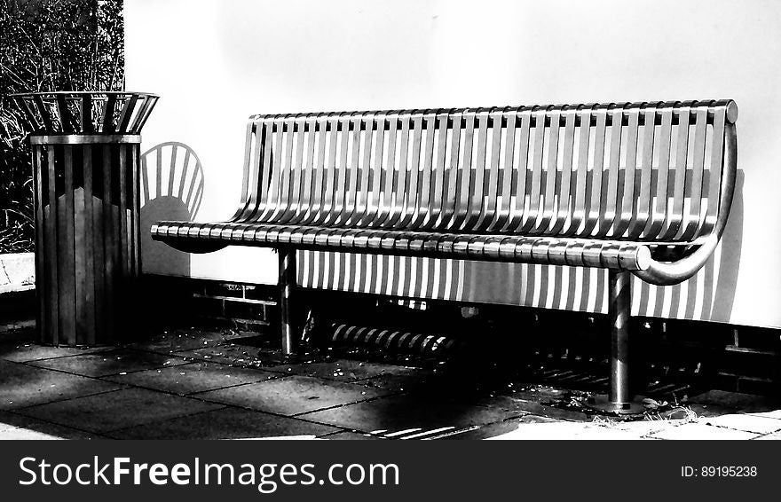 A black and white image of a bench and a trashcan by a wall. A black and white image of a bench and a trashcan by a wall.