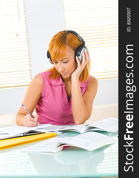 A girl sitting at the table. She's taking notes and listening to music. Front view. A girl sitting at the table. She's taking notes and listening to music. Front view.