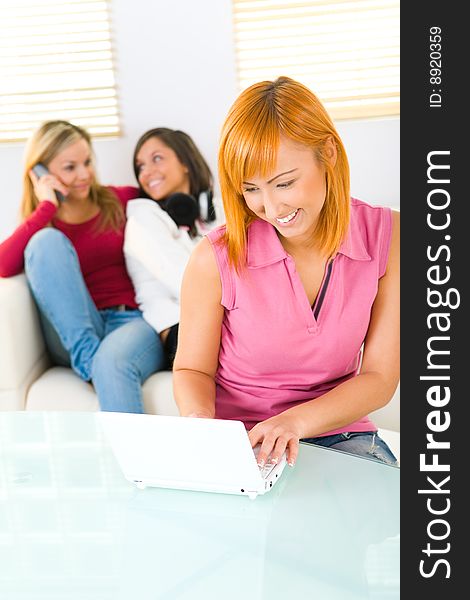 Three young women. One of them sitting at table and doing sometning on laptop. Focused on red-haired girl. Front view. Three young women. One of them sitting at table and doing sometning on laptop. Focused on red-haired girl. Front view.