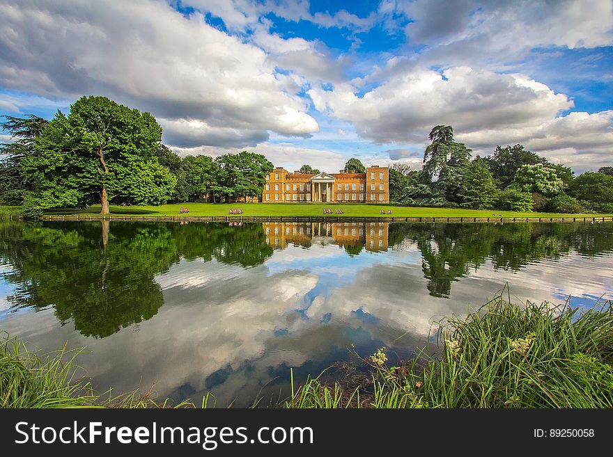 Brown and White House on Green Grass Lawn Surrounded by Trees in Front of Lake Under Blue and White Cloudy Skyt