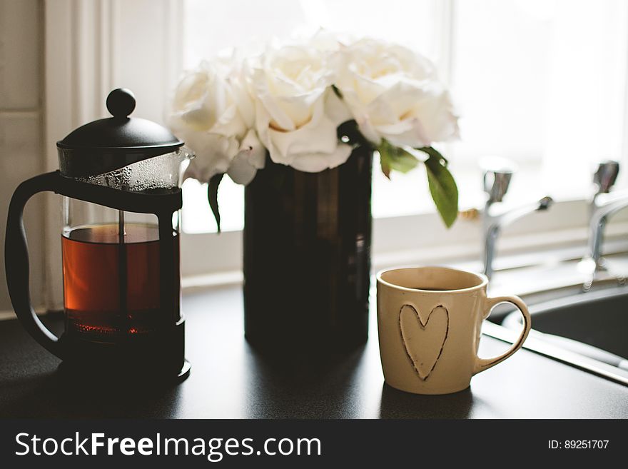 A French press coffee or tea maker and cup with flowers on a kitchen table.