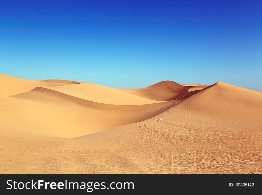 Sand dunes in the desert with vehicle tracks possibly indicating a tourist adventure or safari, cloudless blue sky.