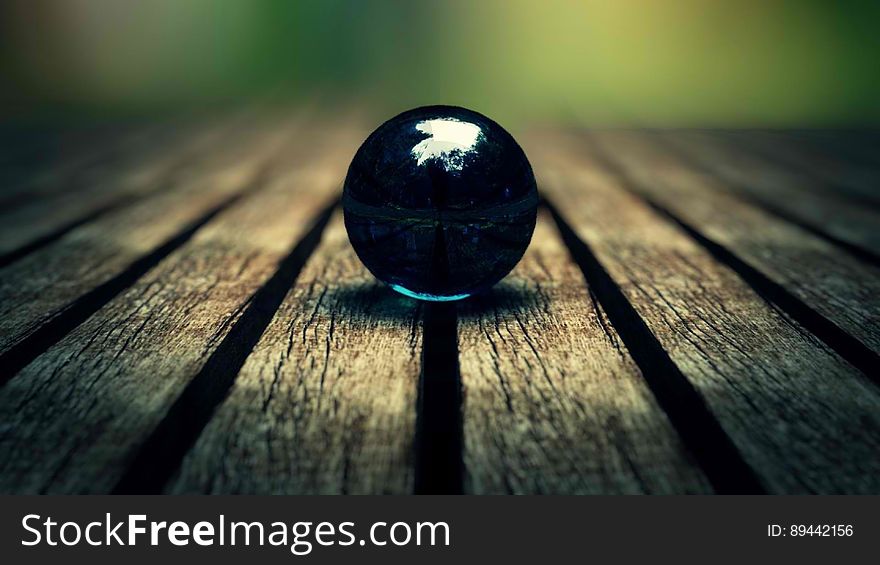 Selective focus on a black marble supported on a series of old wooden planks (table top perhaps) with spaces between each one, blurred background. Selective focus on a black marble supported on a series of old wooden planks (table top perhaps) with spaces between each one, blurred background.