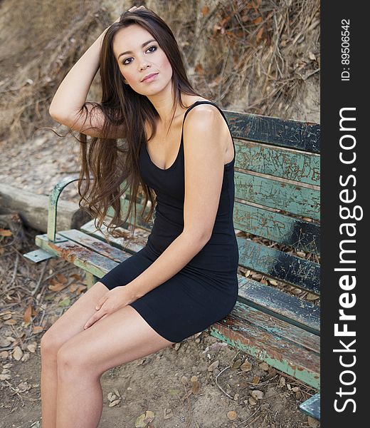 Portrait of young woman with long black hair wearing black dress sitting outdoors on park bench. Portrait of young woman with long black hair wearing black dress sitting outdoors on park bench.