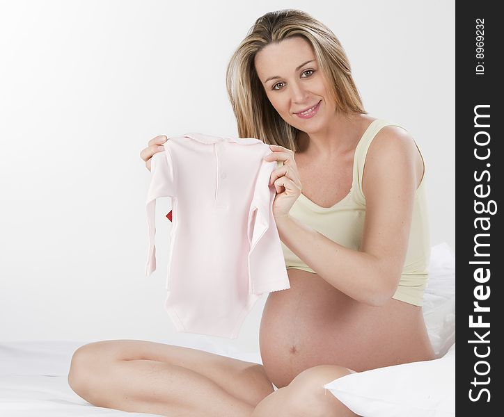 Beautiful Pregnant Young Woman Holding Up New Baby Clothes, Waiting For Her Baby To Be Born. Beautiful Pregnant Young Woman Holding Up New Baby Clothes, Waiting For Her Baby To Be Born