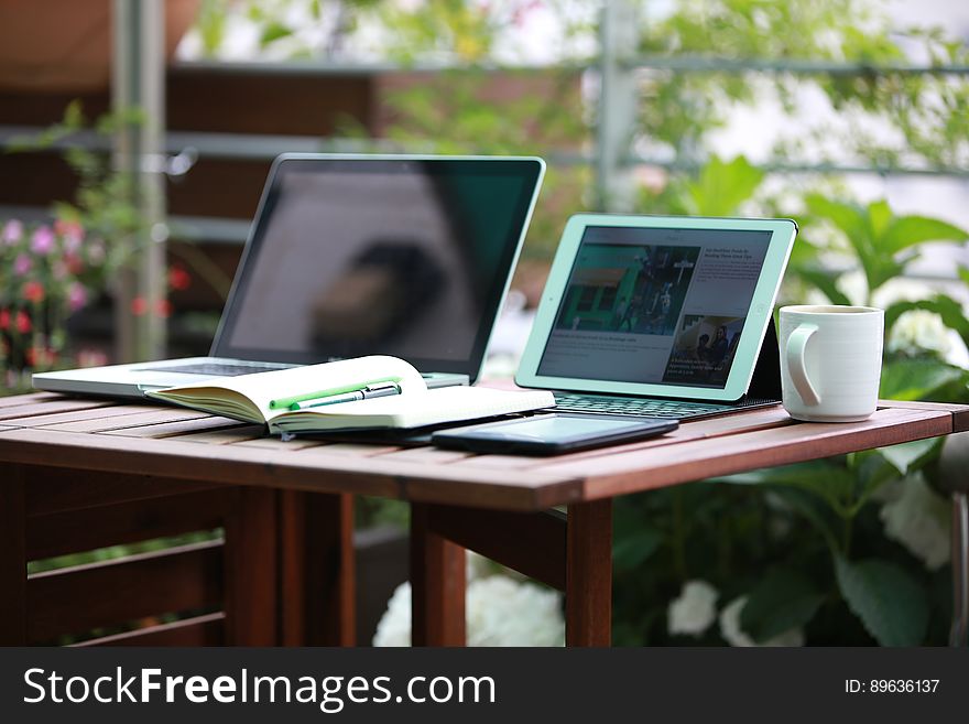 Laptop computers and coffee mug with open notebook on wooden table in sunny garden. Laptop computers and coffee mug with open notebook on wooden table in sunny garden.