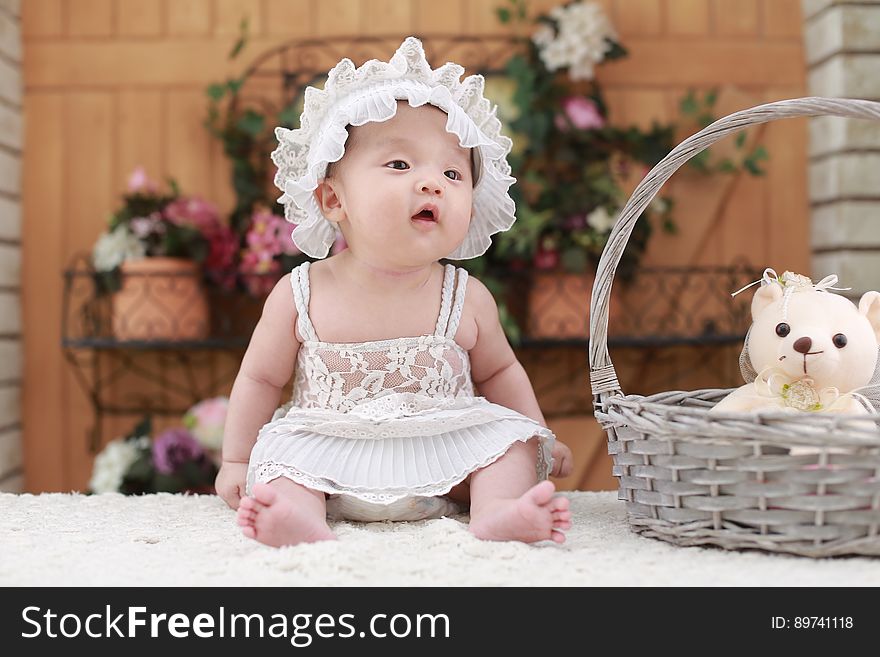 A young baby sitting on table wearing a white dress and a hat. A young baby sitting on table wearing a white dress and a hat.