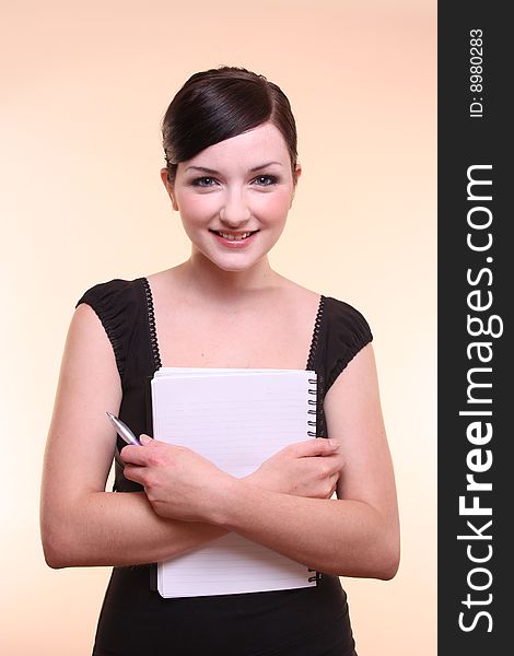 A smiling young professional looking woman holding a notepad in front of a peach background. A smiling young professional looking woman holding a notepad in front of a peach background.