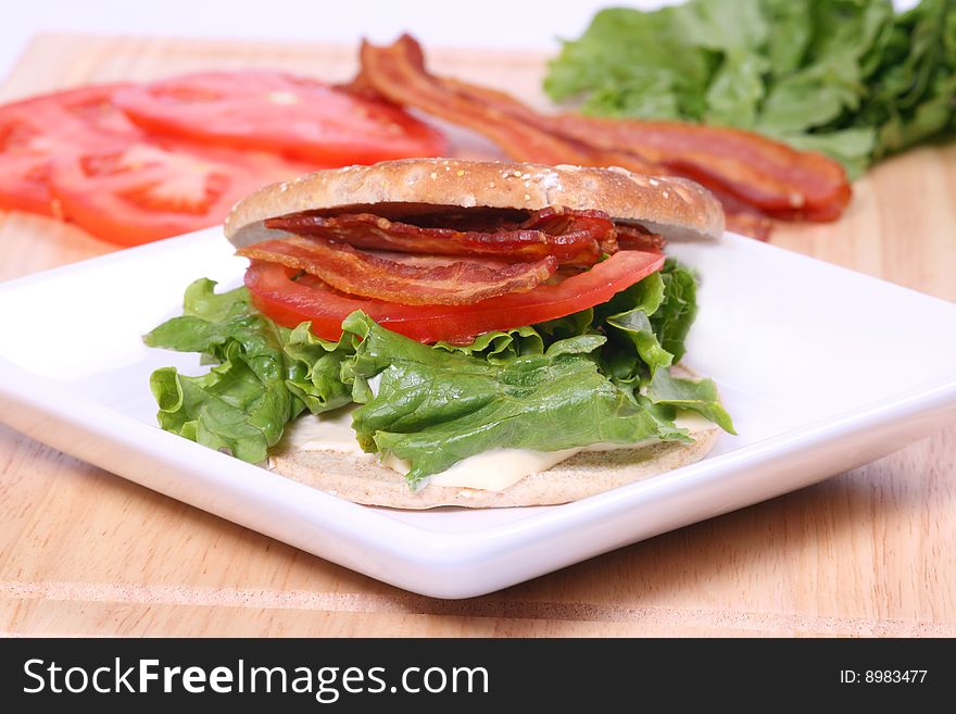 Bacon, lettuce and tomato sandwich with a flat bun on a white plate with ingredients in the background. Bacon, lettuce and tomato sandwich with a flat bun on a white plate with ingredients in the background