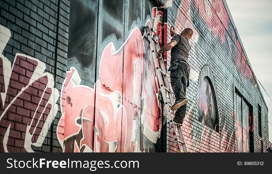Man in Gray Shirt Standing on Gray Steel Ladder Painting Black White and Red Graffiti on Concrete Wall Outdoors
