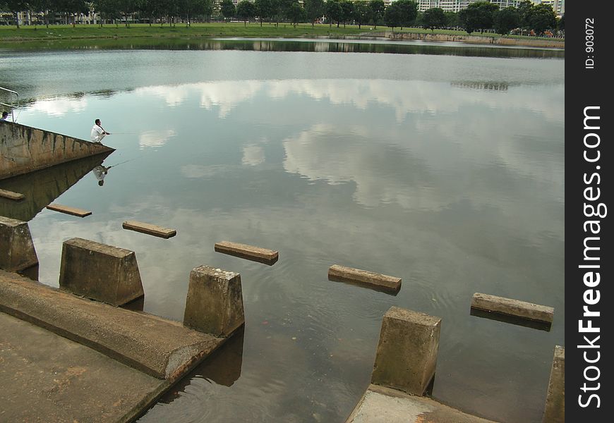 Taken in yishun, singapore. Man is squating on concrete slope fishing in a man made reservoir. Sky reflected in waters.  . Taken in yishun, singapore. Man is squating on concrete slope fishing in a man made reservoir. Sky reflected in waters.