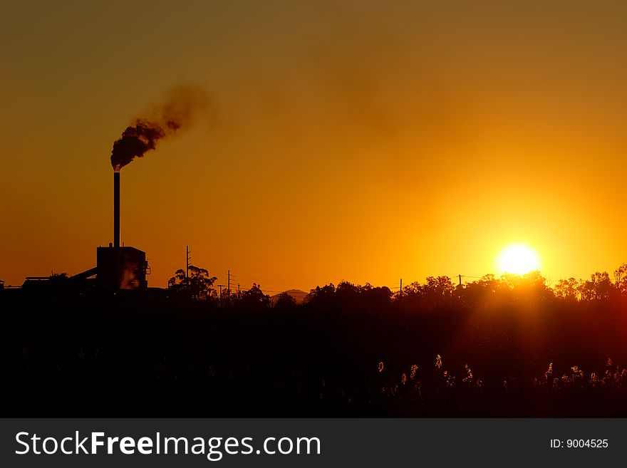 An industrial smoke stack ejects smoke against an clear orange sunset while foreground plants reach for the last rays of sunshine. An industrial smoke stack ejects smoke against an clear orange sunset while foreground plants reach for the last rays of sunshine