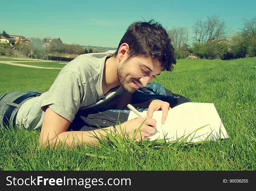 Young dark haired man lying almost prone in a green grassy field but writing (or drawing) on a sheet of white paper, blue sky. Young dark haired man lying almost prone in a green grassy field but writing (or drawing) on a sheet of white paper, blue sky.