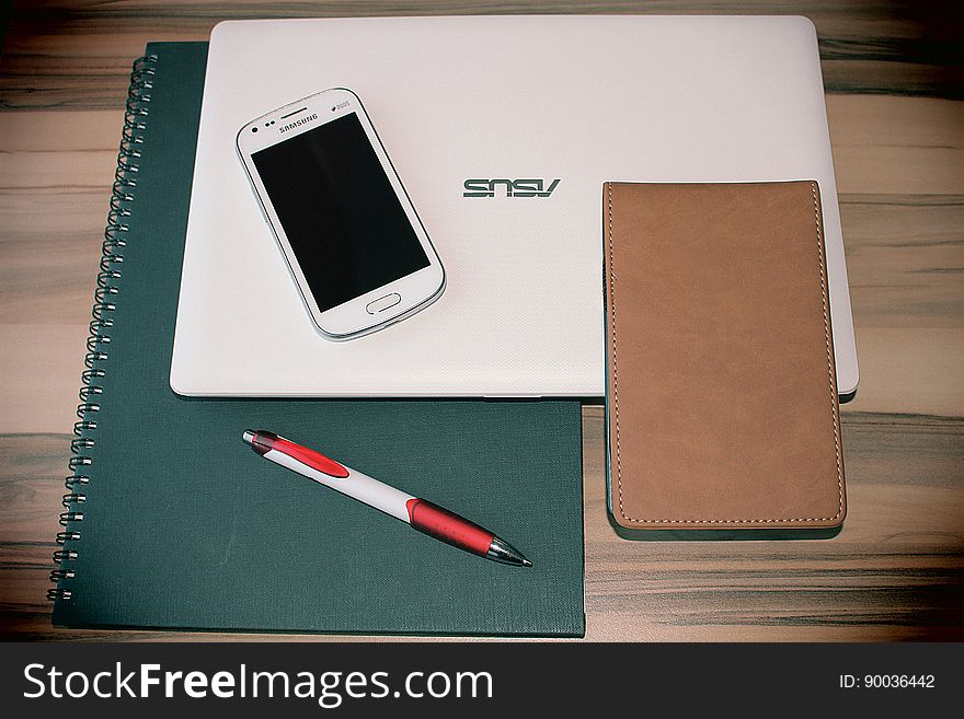 Spiral notebook, laptop computer, mobile phone and leather case and red and white pen on a wooden table. Spiral notebook, laptop computer, mobile phone and leather case and red and white pen on a wooden table.
