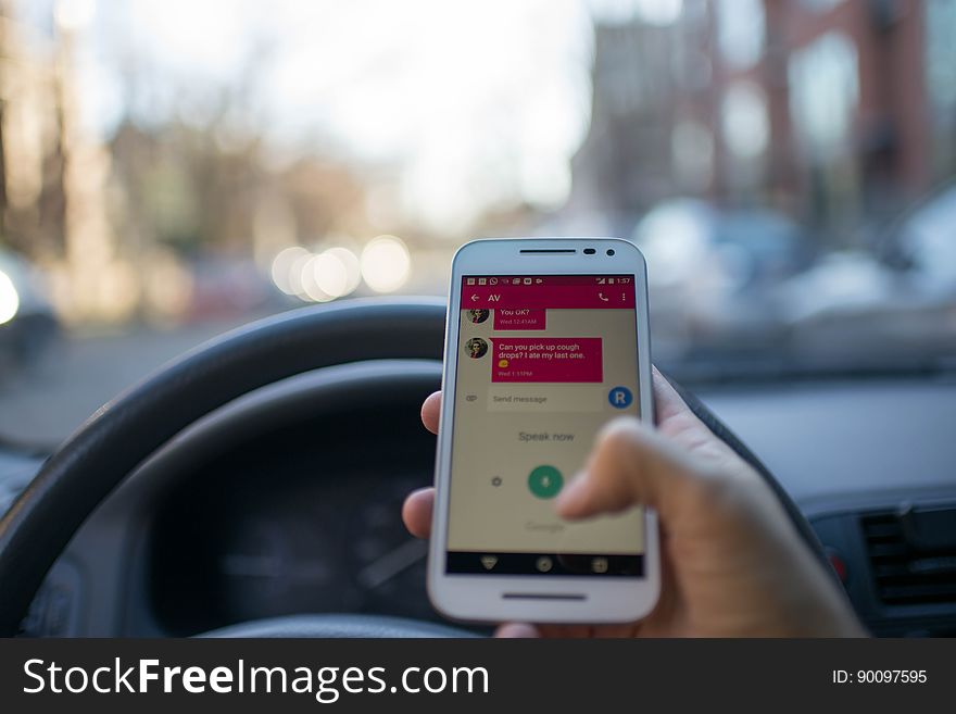A person behind the steering wheel of a car with a smartphone in hand. A person behind the steering wheel of a car with a smartphone in hand.