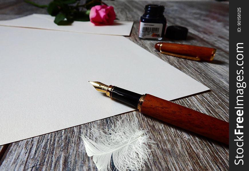 Fountain pen on blank sheet of paper with jar of ink, rose and feather on rustic wooden tabletop. Fountain pen on blank sheet of paper with jar of ink, rose and feather on rustic wooden tabletop.