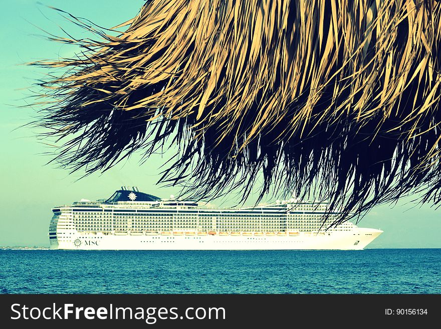 View under thatched beach umbrella of cruise ship in blue waters on sunny day. View under thatched beach umbrella of cruise ship in blue waters on sunny day.