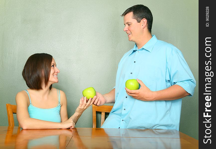 Beautiful young male caucasian offering green apple to his girlfriend or wife on kitchen table. Beautiful young male caucasian offering green apple to his girlfriend or wife on kitchen table