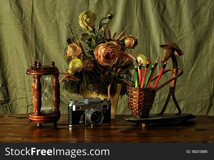 Still life on table top with timer, vase of (paper) roses, basket of colored pencils and retro film camera green curtain background. Still life on table top with timer, vase of (paper) roses, basket of colored pencils and retro film camera green curtain background.