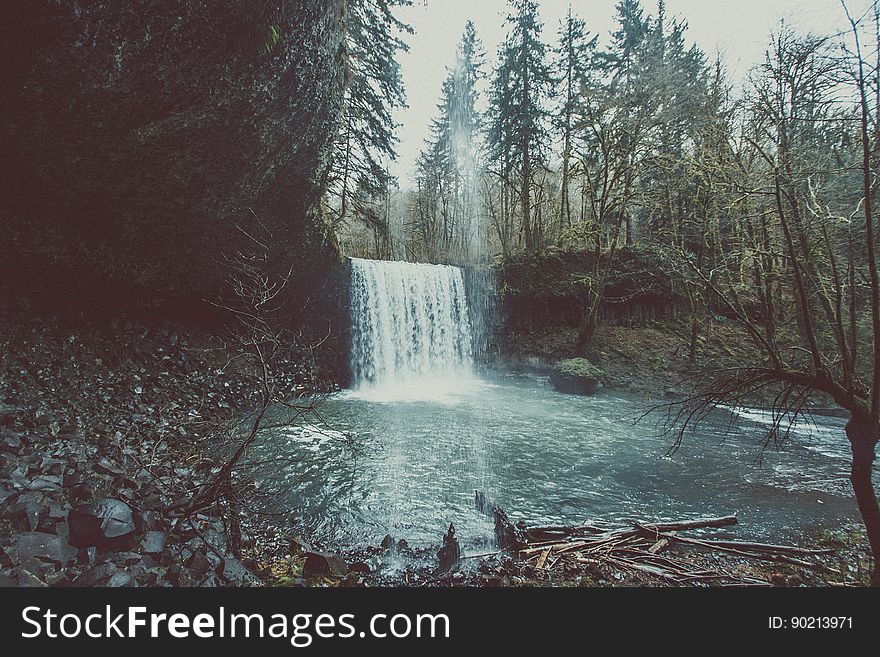 Waterfall in fir forest with relatively modest drop into a pool with stones, rocks and debris carried down by flooding, gray sky. Waterfall in fir forest with relatively modest drop into a pool with stones, rocks and debris carried down by flooding, gray sky.