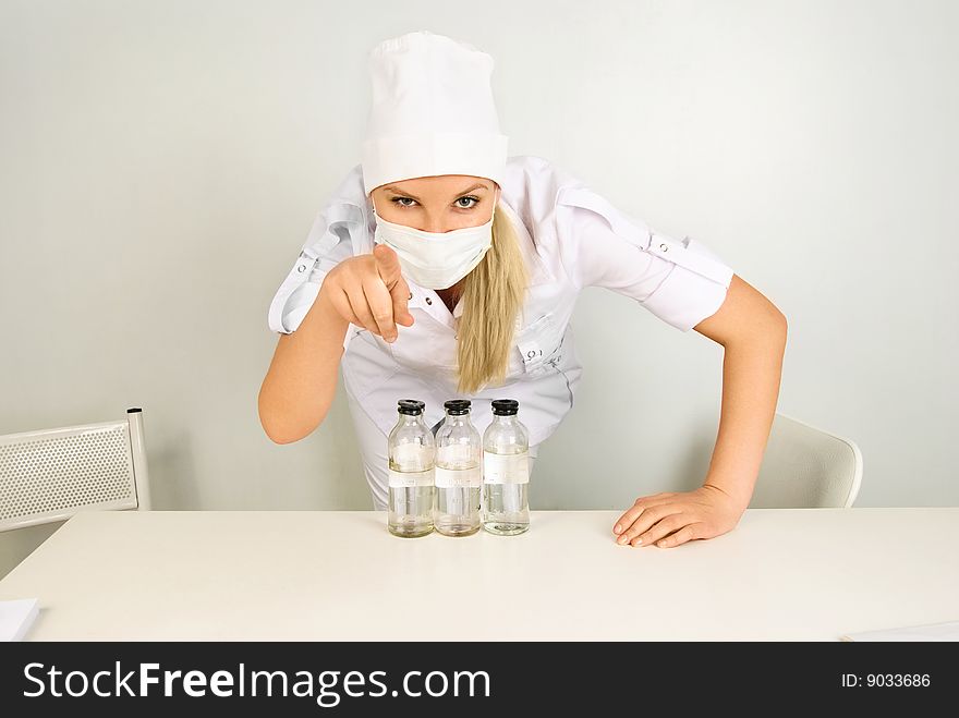 Portrait of a young doctor with medications sitting by the table in her office and pointing at us with a finger. Portrait of a young doctor with medications sitting by the table in her office and pointing at us with a finger