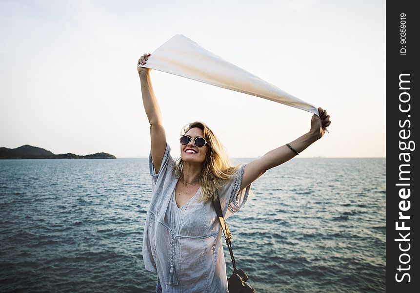 Smiling blond woman in pale blue dress with her arms outstretched holding a piece of white material, background of sea and waves. Smiling blond woman in pale blue dress with her arms outstretched holding a piece of white material, background of sea and waves.