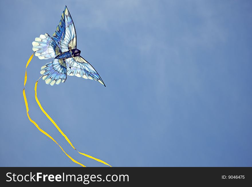 Blue toned-colored kite flies against a blue sky background with white clouds, yellow streams trailing in a circular arc. Blue toned-colored kite flies against a blue sky background with white clouds, yellow streams trailing in a circular arc