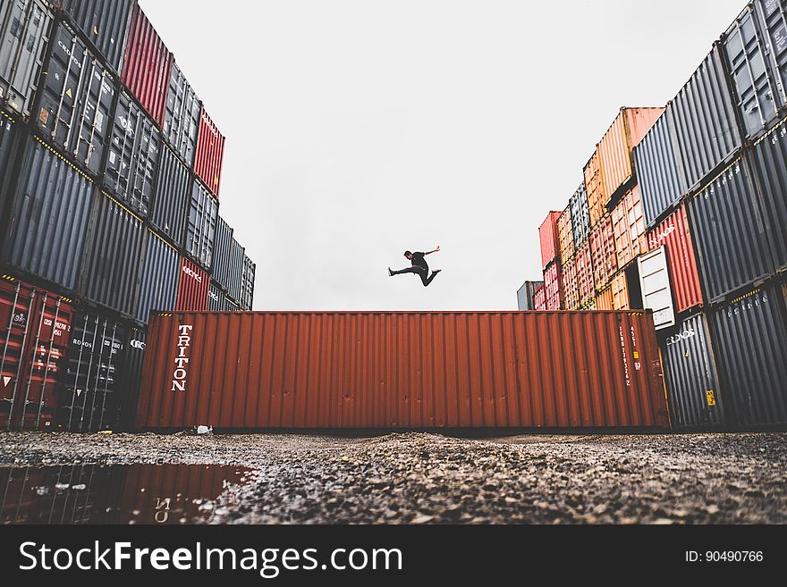 Athlete jumping high over one of a collection of containers used for export and import of goods by ship or rail, pale gray sky background. Athlete jumping high over one of a collection of containers used for export and import of goods by ship or rail, pale gray sky background.