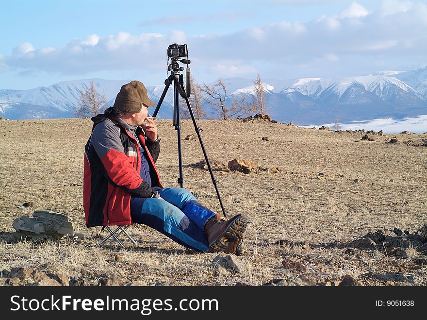 Old man sitting and in the mountains with camera standing on tripod. Old man sitting and in the mountains with camera standing on tripod