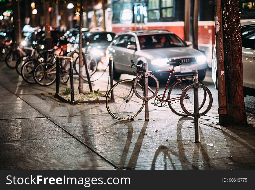 Bicycles parked at the side of a street with cars passing close by, their headlights casting long shadows over the wet sidewalk. Bicycles parked at the side of a street with cars passing close by, their headlights casting long shadows over the wet sidewalk.