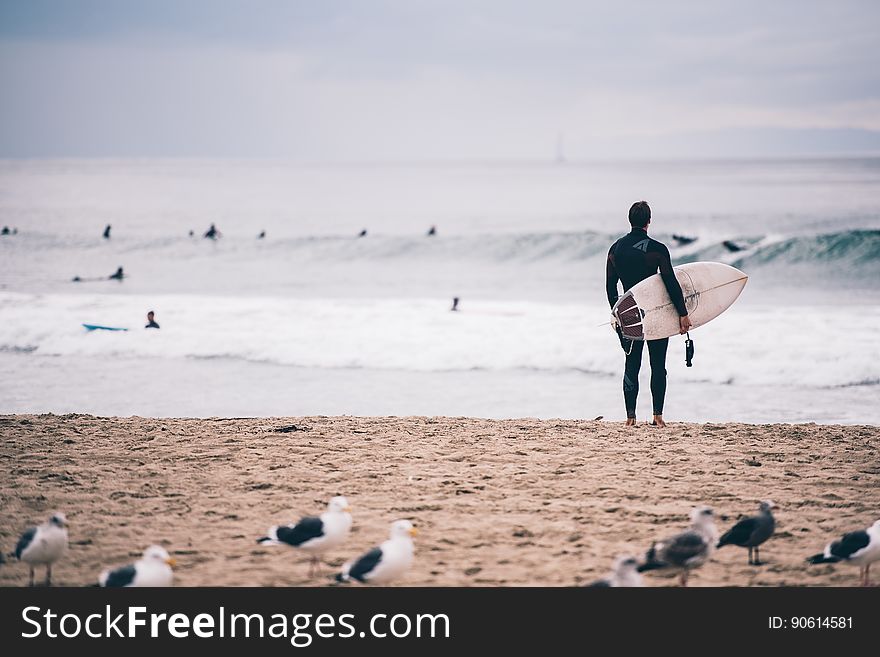 Man, possibly wearing wet suit, holding a white surf board and standing on the sandy beach assessing the waves. Man, possibly wearing wet suit, holding a white surf board and standing on the sandy beach assessing the waves.