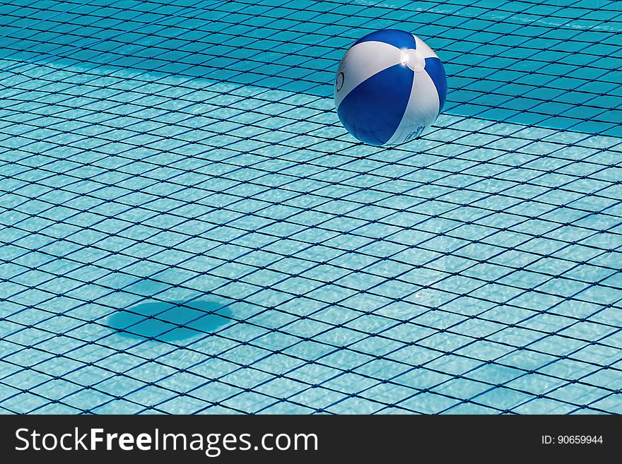 A blue and white beach ball floating on the surface of a swimming pool with tiled bottom. A shadow of the ball is cast on the bottom of the pool. A blue and white beach ball floating on the surface of a swimming pool with tiled bottom. A shadow of the ball is cast on the bottom of the pool.