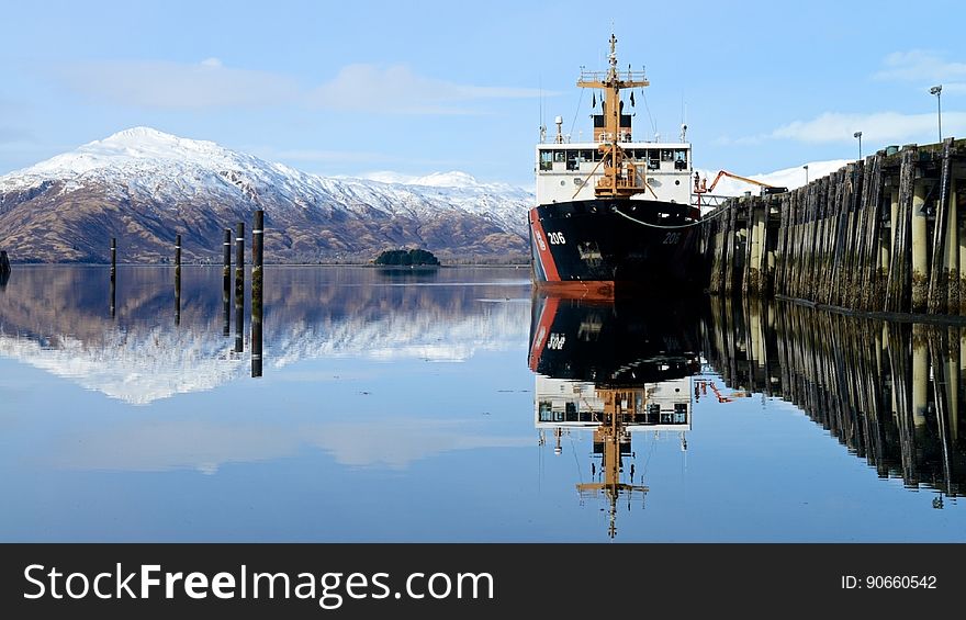 Ship in dock reflecting in clear blue waters with snow capped mountains in horizon with blue skies on sunny day.