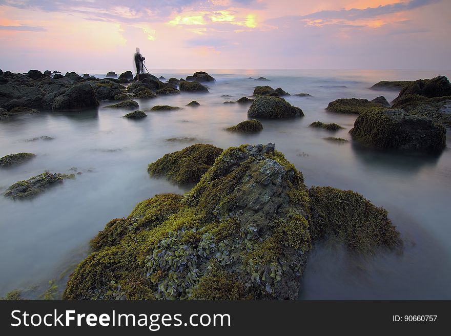 Blur of water over rocky shores with silhouette of standing person against sunset in cloudy skies. Blur of water over rocky shores with silhouette of standing person against sunset in cloudy skies.