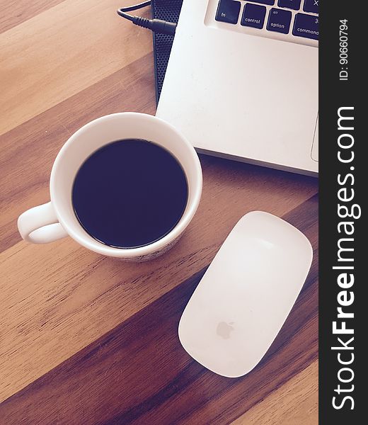 Overhead view of white china cup of coffee nest to laptop computer and Apple mouse on wooden tabletop. Overhead view of white china cup of coffee nest to laptop computer and Apple mouse on wooden tabletop.