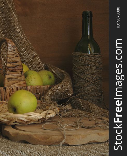 Green apples in a basket, a bottle, a chopping board against a homespun drapery. Green apples in a basket, a bottle, a chopping board against a homespun drapery