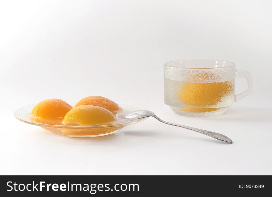 Segments of tinned peaches on a glass saucer with a dessertspoon and a cup with a peach and a syrup. Segments of tinned peaches on a glass saucer with a dessertspoon and a cup with a peach and a syrup