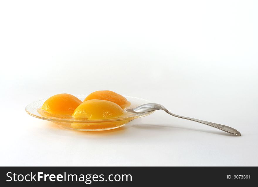 Segments of tinned peaches on a glass saucer with a dessertspoon on a white background. Segments of tinned peaches on a glass saucer with a dessertspoon on a white background