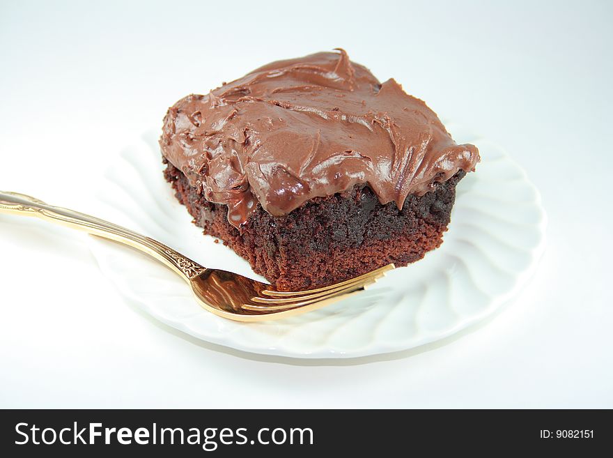 A frosted chocolate brownie on a white plate with a gold colored fork. A frosted chocolate brownie on a white plate with a gold colored fork.