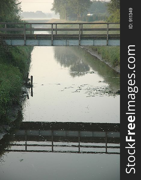 A bridge and its reflection crossing a still creek in the mist. A bridge and its reflection crossing a still creek in the mist.