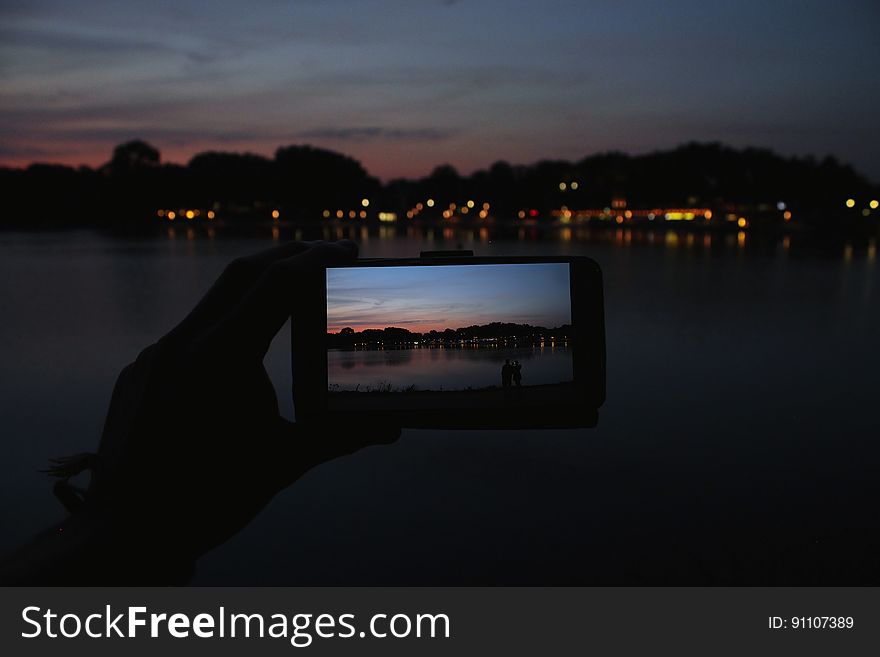 Hand holding smartphone with image of coastal sunset against landscape. Hand holding smartphone with image of coastal sunset against landscape.