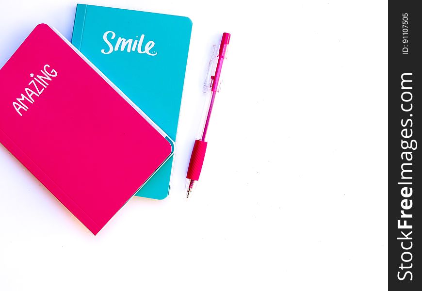 Two small notebooks, a red one marked "Amazing" and a blue one marked "Smile" alongside is a red pen, white background. Two small notebooks, a red one marked "Amazing" and a blue one marked "Smile" alongside is a red pen, white background.