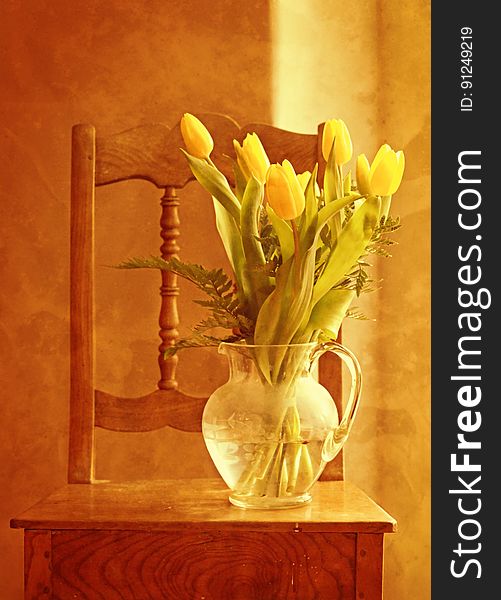 Glass pitcher vase with yellow tulips on small wooden table with chair in sunlight. Glass pitcher vase with yellow tulips on small wooden table with chair in sunlight.