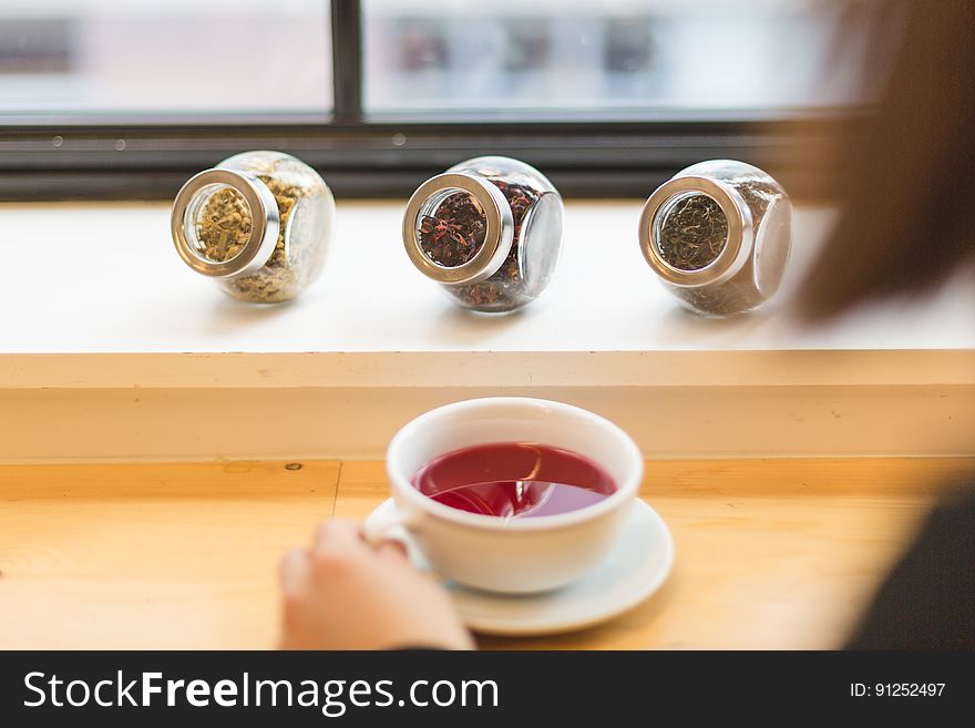 Hand of person holding cup of fruit tea with jars of herbs on ledge by window. Hand of person holding cup of fruit tea with jars of herbs on ledge by window.